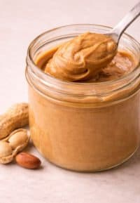 Jar of creamy peanut butter with spoon and whole peanuts, the smooth and nutty base layer ingredient in weScotchoo Scotcheroo Bars.
