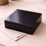 Glossy black Scotcheroo gift box closed and ready for shipping, displayed on a neutral linen surface with soft natural lighting.