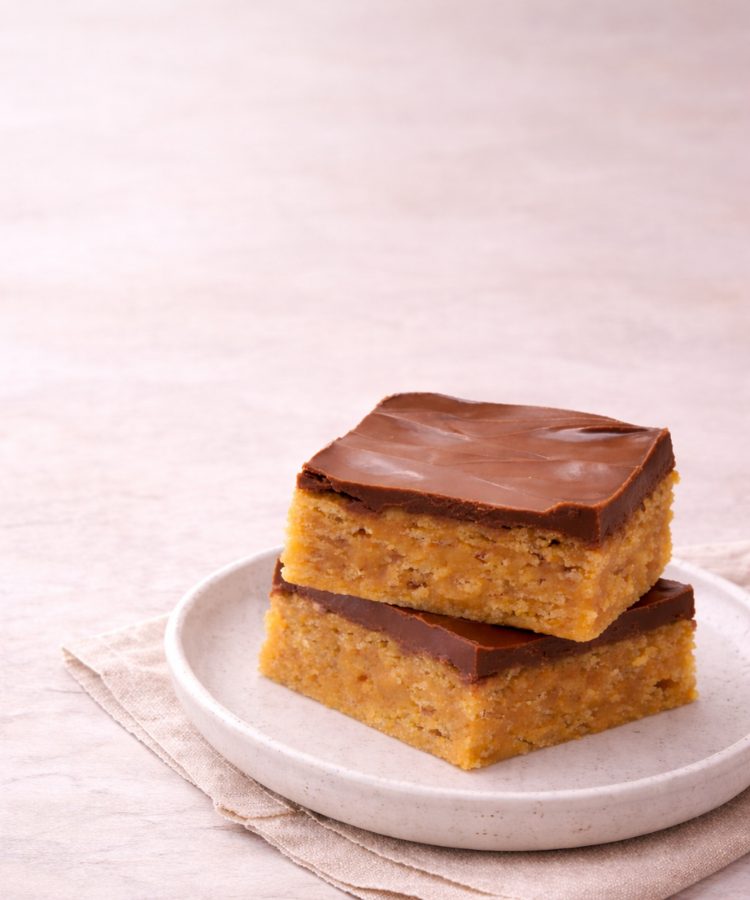 Stacked Scotcheroo Bars with a thick chocolate topping and chewy peanut butter cereal base on a ceramic plate against a soft beige background. {{brizy_dc_image_alt imageSrc=