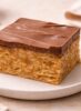 Handcrafted Scotcheroo bar with thick chocolate and butterscotch topping on a neutral ceramic plate, small-batch peanut butter rice cereal dessert.