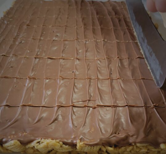 Pan of Scotcheroos being sliced into bars using the homemade scotcheroos recipe by weScotchoo.com – soft, chewy, and topped with chocolate and butterscotch.